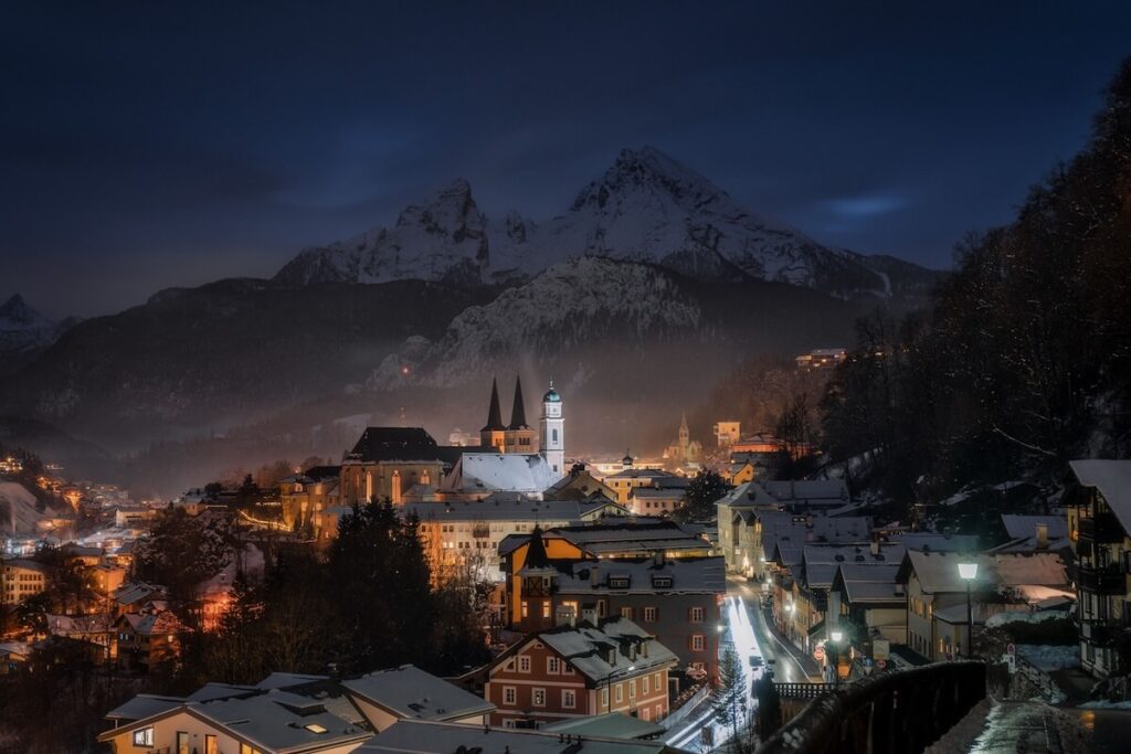 A picturesque winter night view of Berchtesgaden, Germany, with snow-covered rooftops and majestic mountains in the background.