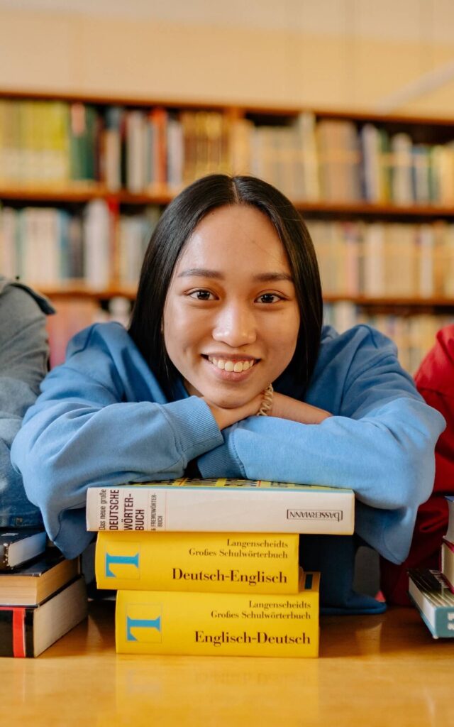 student in a library leaning on a stack of German-English books