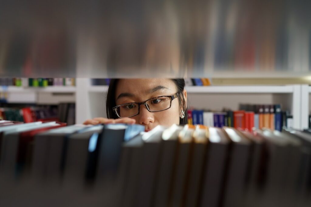 girl wearing glasses standing between bookshelves in university library, picking up book from shelf