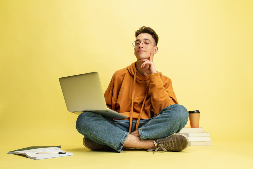 student in glasses sits on floor with laptop isolated on yellow studio background