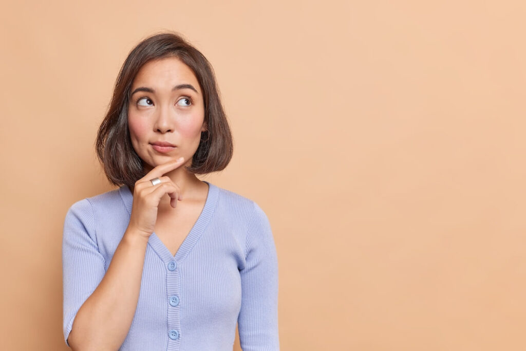 Thoughtful Asian woman keeps hand on chin looks pensively above