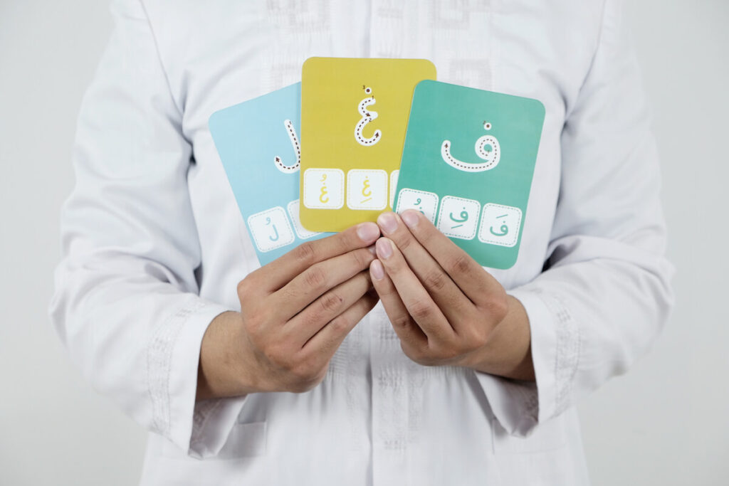 Someone wearing a white Muslim shirt or Koko shirt is holding three Arabic letters cards on his chest