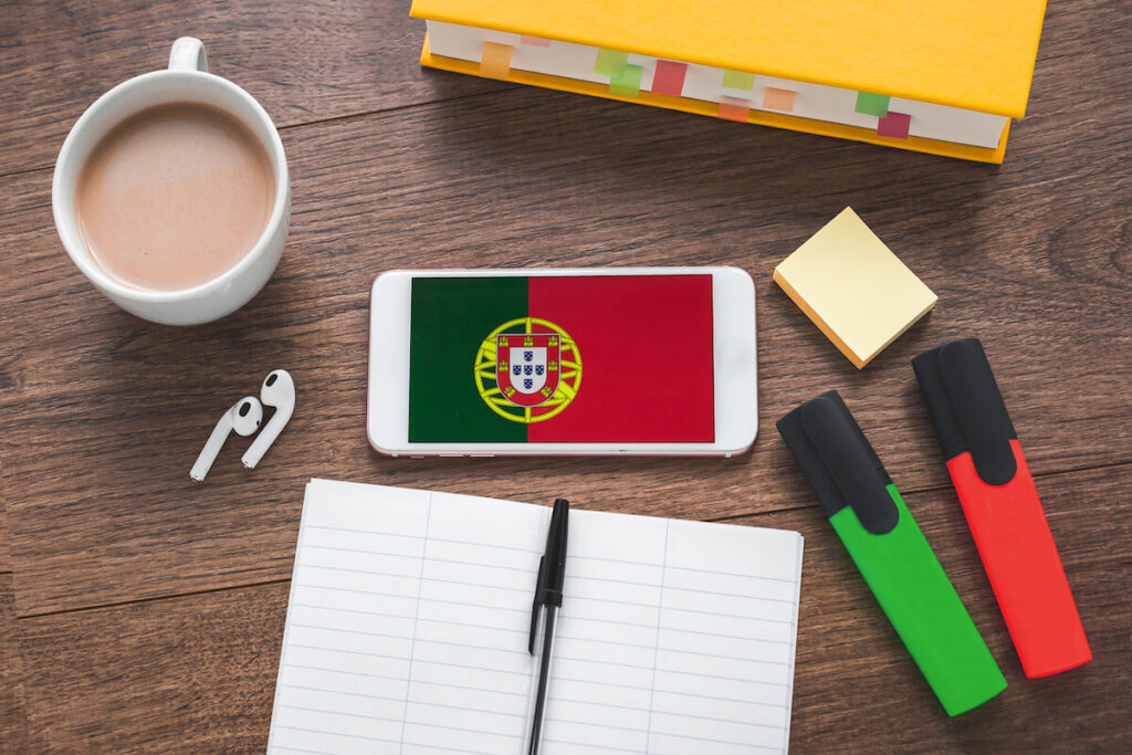 smartphone with portuguese flag and coat of arms, wireless headphones, notepad, textbook and cup of coffee on a brown wooden table