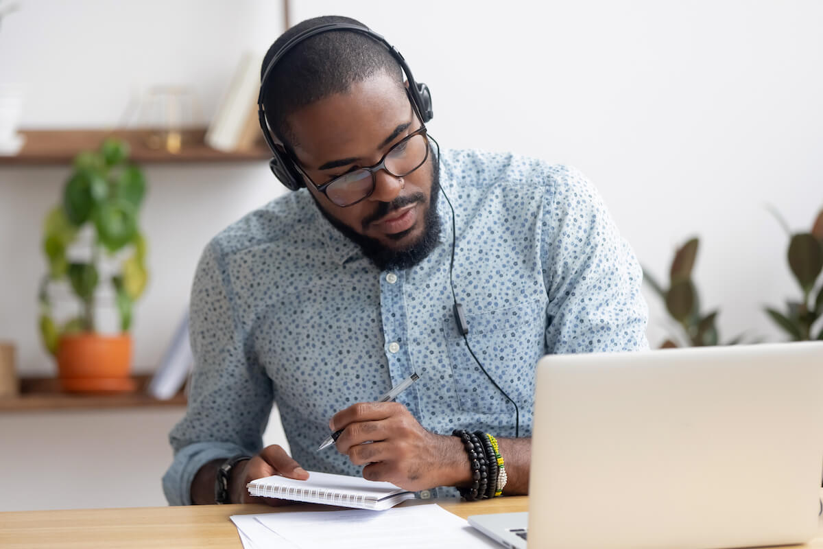man taking notes while listening on headphones and looking at laptop