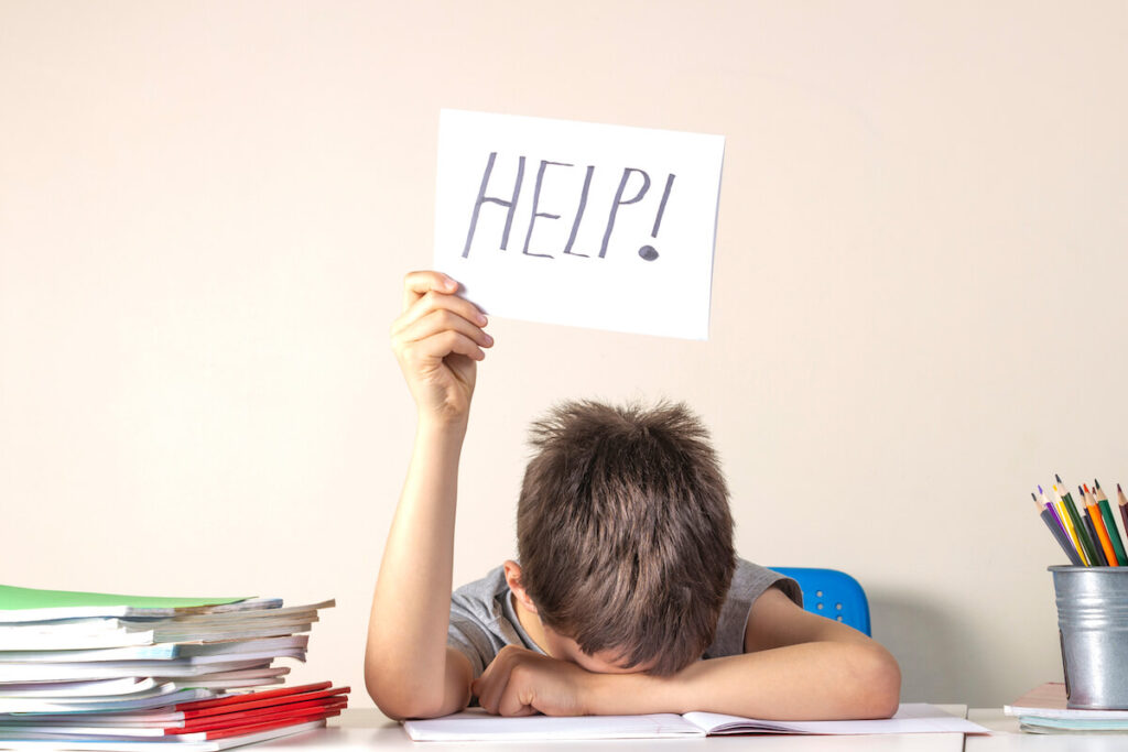 frustrated boy sitting at the table with many books and holding paper with word Help