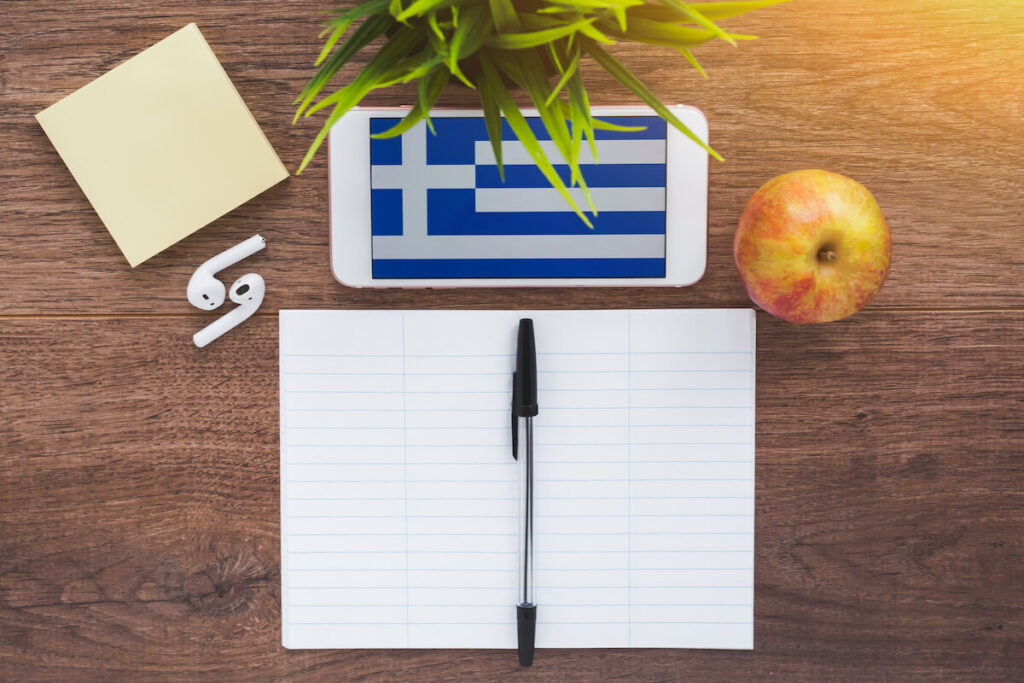 Greece flag, smartphone, wireless headphones, a notebook for writing foreign words on a working wooden table, the concept of learning a foreign language