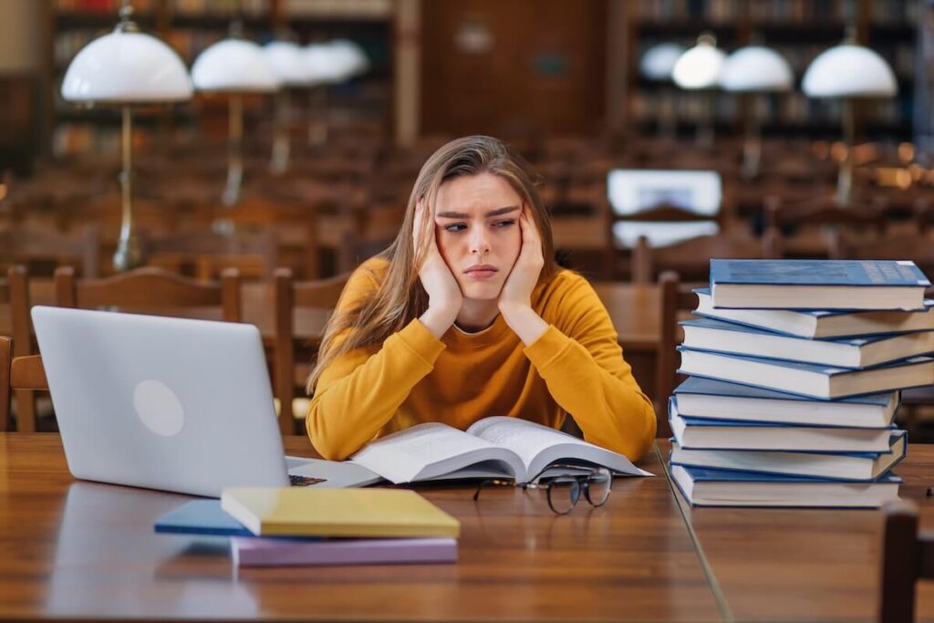 female student stressed, pile of books and laptop on desk